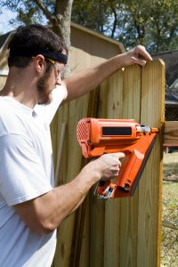 28065930 - man wearing safety glasses uses a portable nail gun to attach wood pickets to the rail as he builds a privacy fence in the backyard.